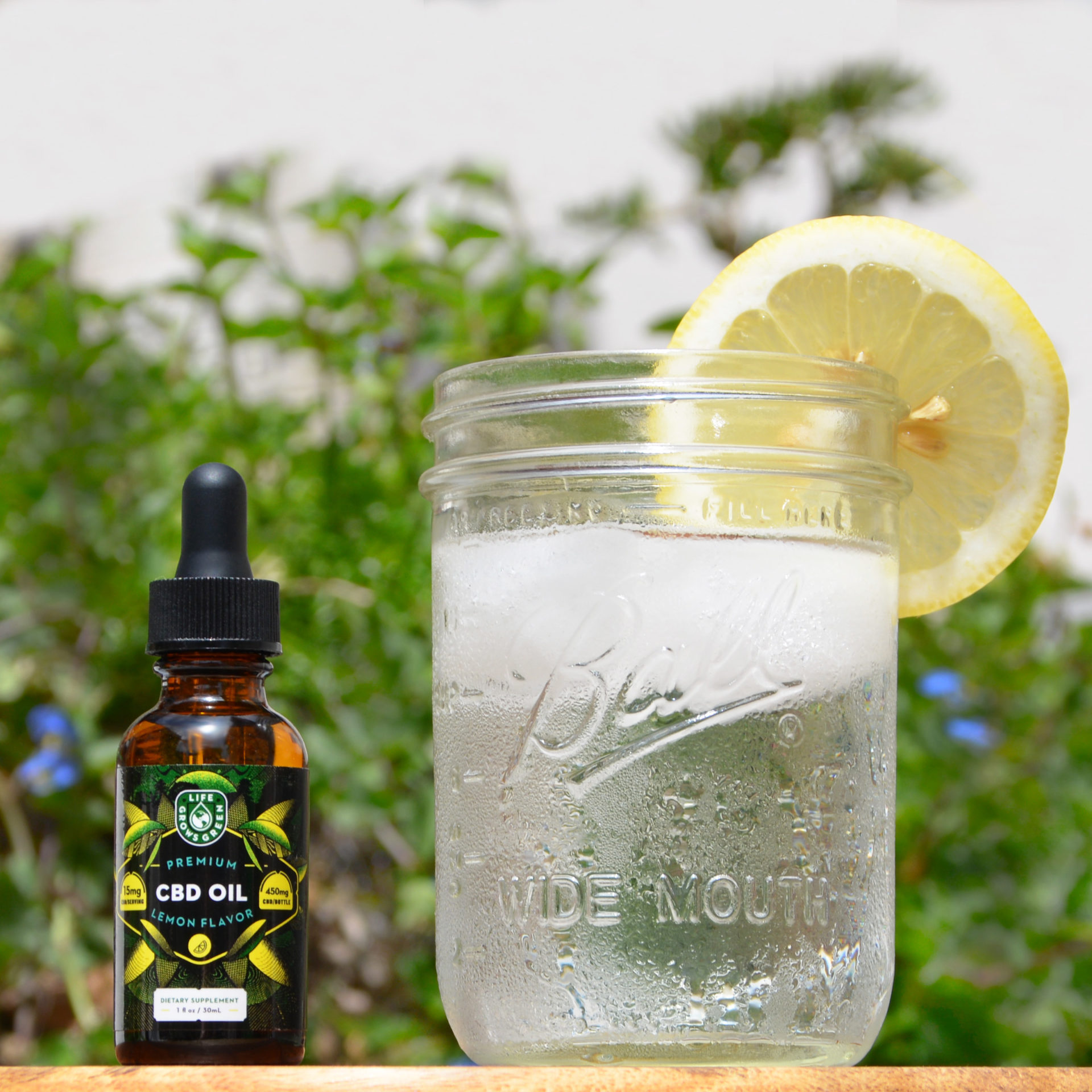 A bottle of lemon flavored Life Grows Green cbd oil next to a glass of lemon water.