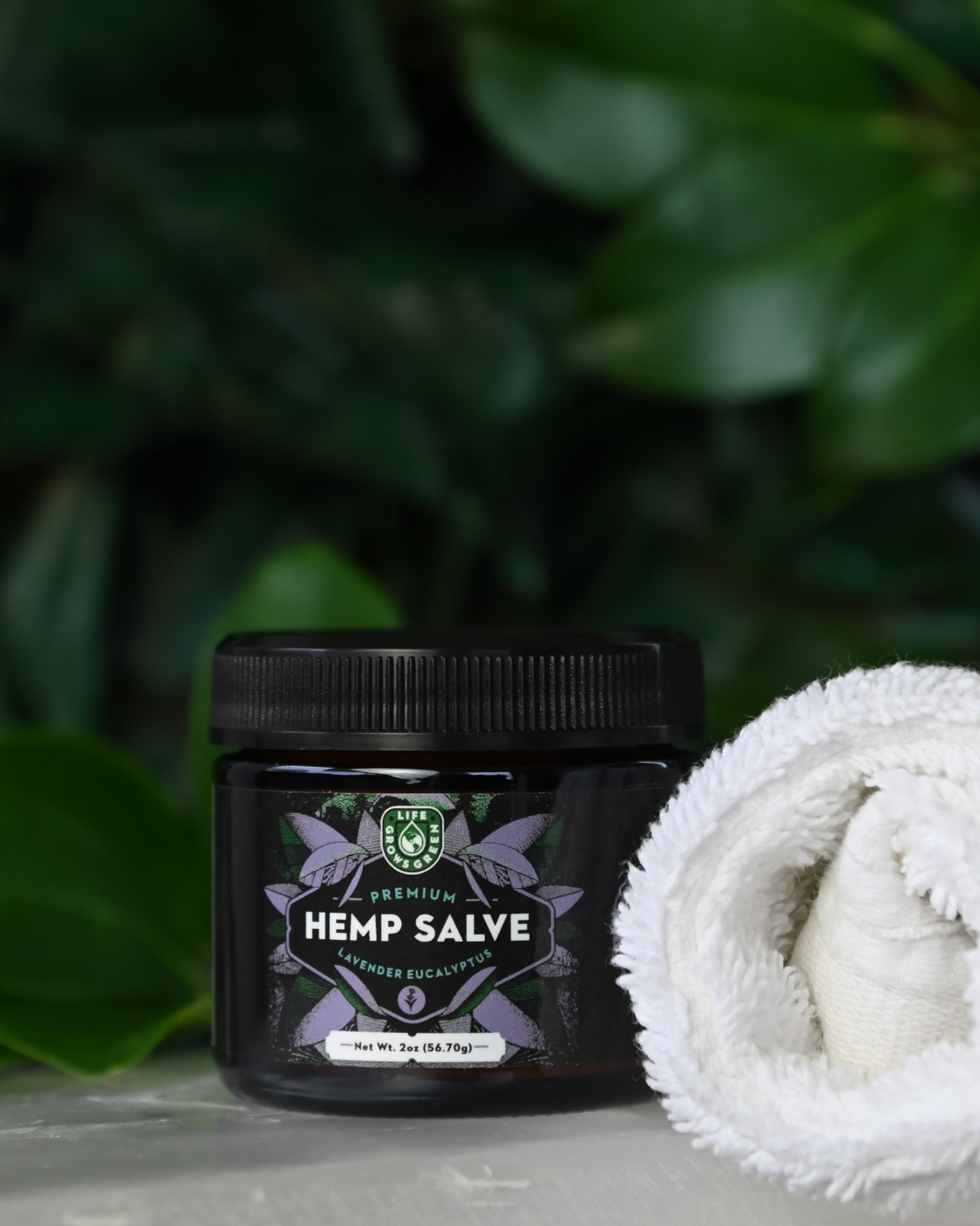 A jar of Lavender Hemp Salve with a white spa towel in front of a green leafy background.