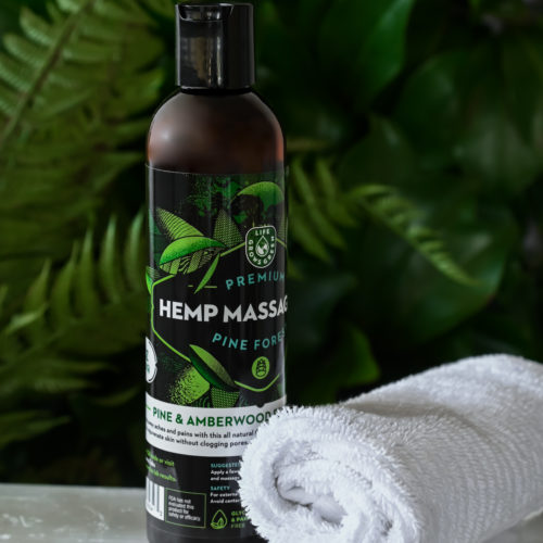 A bottle of hemp massage oil with pine and amberwood next to a spa towel in front of a fern..