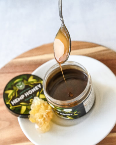 A spoon drizzling hemp honey in to a jar of honey next to a honeycomb.